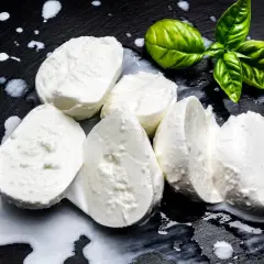 Świeża mozzarella na czarnym tle, z kilkoma listkami bazylii. Czy mozzarella jest kaloryczna? To zależy od jej rodzaju, ale zazwyczaj jest umiarkowanie kaloryczna.