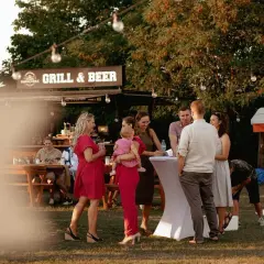 Ludzie na imprezie plenerowej przy grillu i piwie, część osób stoi przy stołach, część siedzi.
