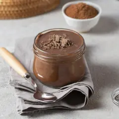 Słoik z czekoladowym puddingiem proteinowym, posypanym wiórkami czekolady, obok łyżeczki i miseczki z nasionami chia.