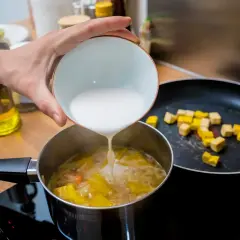 Dodawanie mleczka kokosowego do zupy, by zagęścić ją bez mąki. Na patelni smażą się kostki tofu.