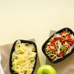Dwa pojemniki z jedzeniem: makaron farfalle i sałatka z groszku, kapusty i papryki. Obok jabłko i łyżka.