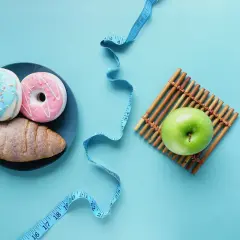 Dwa pączki i rogalik na talerzu obok zielonego jabłka i miarki krawieckiej. Wybór między słodyczami a zdrowiem, między kj a kcal.