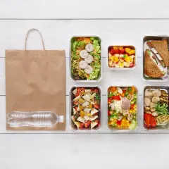 Co do grama catering: pięć pudełek z jedzeniem, torba i butelka wody na białym drewnianym stole.