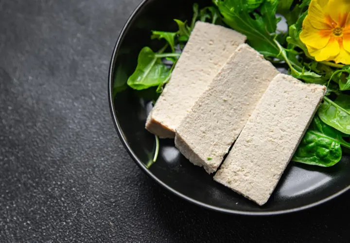 Trzy kawałki tofu na talerzu z zielonymi liśćmi i żółtym kwiatkiem. Czy tofu to nabiał? To pytanie często zadawane przez osoby na diecie roślinnej.