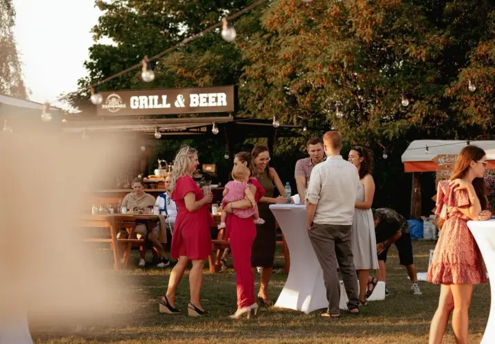 Ludzie na imprezie plenerowej przy grillu i piwie, część osób stoi przy stołach, część siedzi.