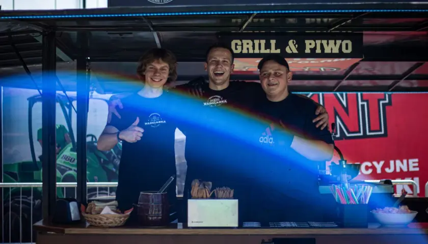 Trzech uśmiechniętych mężczyzn za ladą cateringową z napisem "Grill & Piwo".