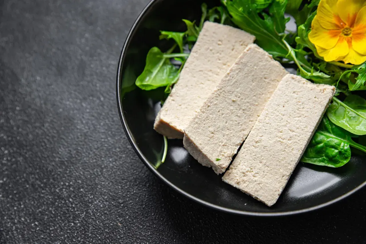 Trzy kawałki tofu na talerzu z zielonymi liśćmi i żółtym kwiatkiem. Czy tofu to nabiał? To pytanie często zadawane przez osoby na diecie roślinnej.