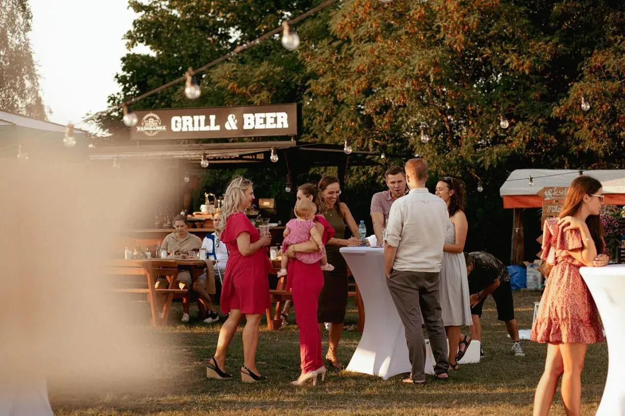 Ludzie na imprezie plenerowej przy grillu i piwie, część osób stoi przy stołach, część siedzi.