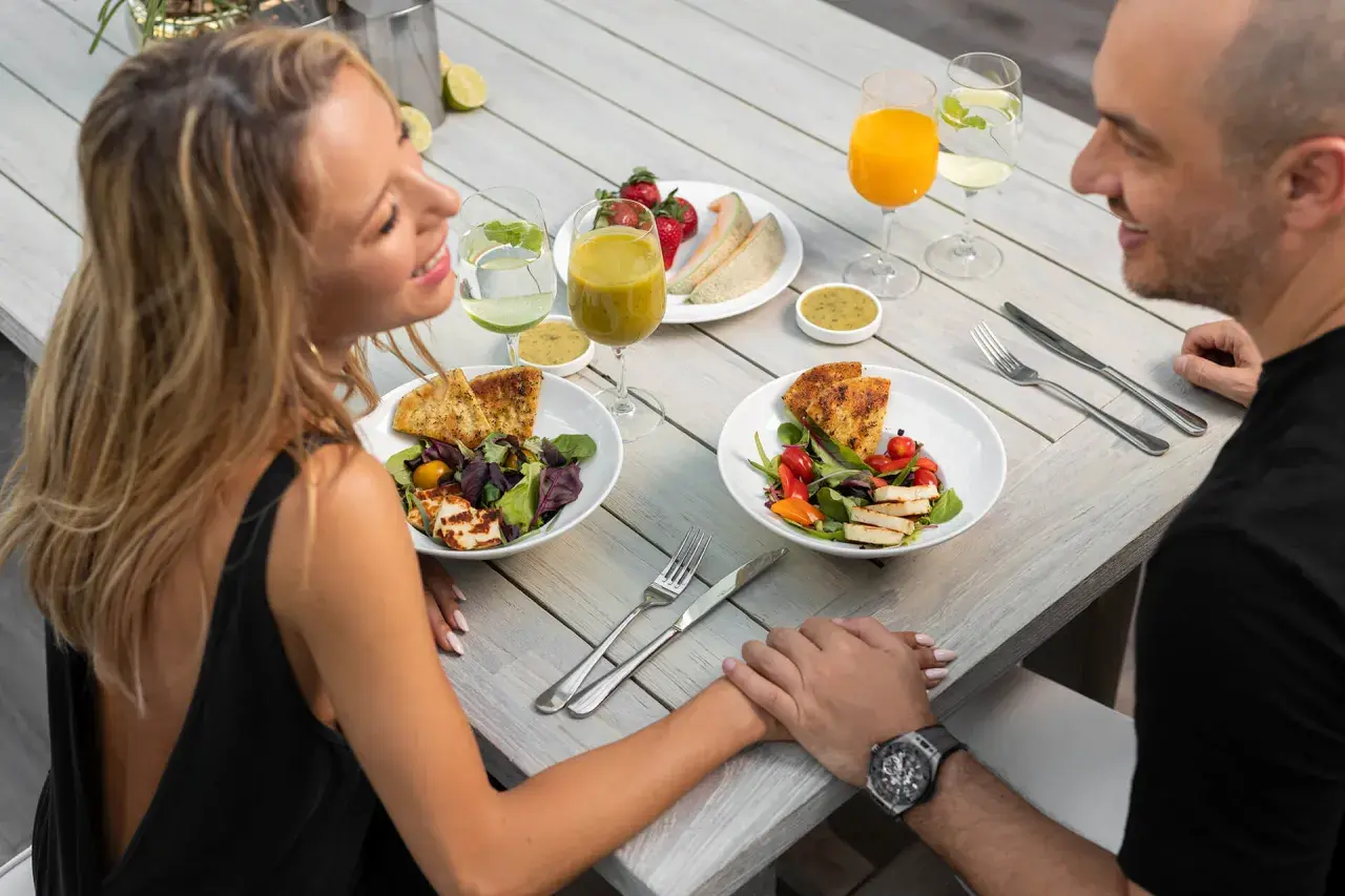 Romantyczny catering dla par. Uśmiechnięta para trzyma się za ręce przy stole z pysznymi sałatkami, owocami i napojami.