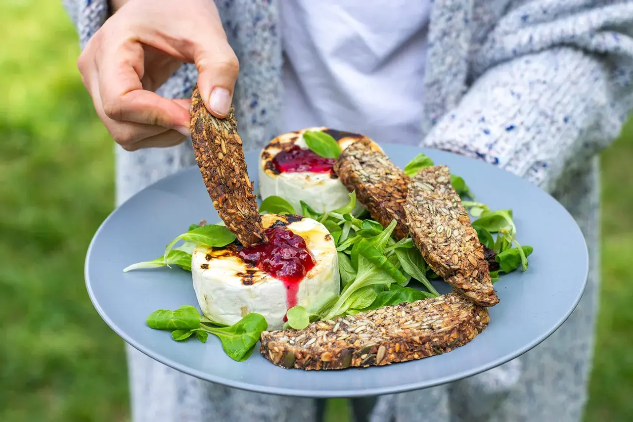 Ręka zanurza chrupiący sucharek w grillowanym serze z konfiturą. Czy takie sucharki są kaloryczne? Zależy od składu, ale te pełne ziaren wyglądają zdrowo!