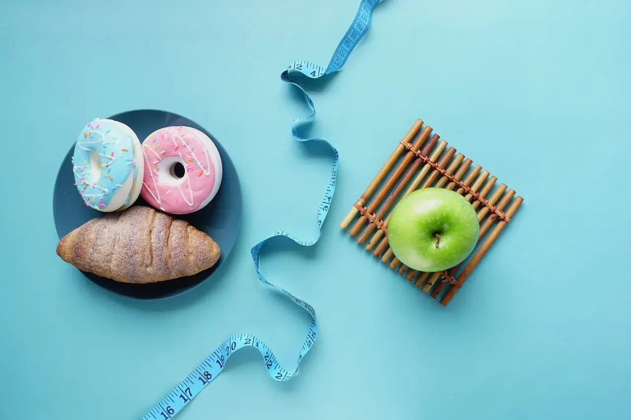 Dwa pączki i rogalik na talerzu obok zielonego jabłka i miarki krawieckiej. Wybór między słodyczami a zdrowiem, między kj a kcal.