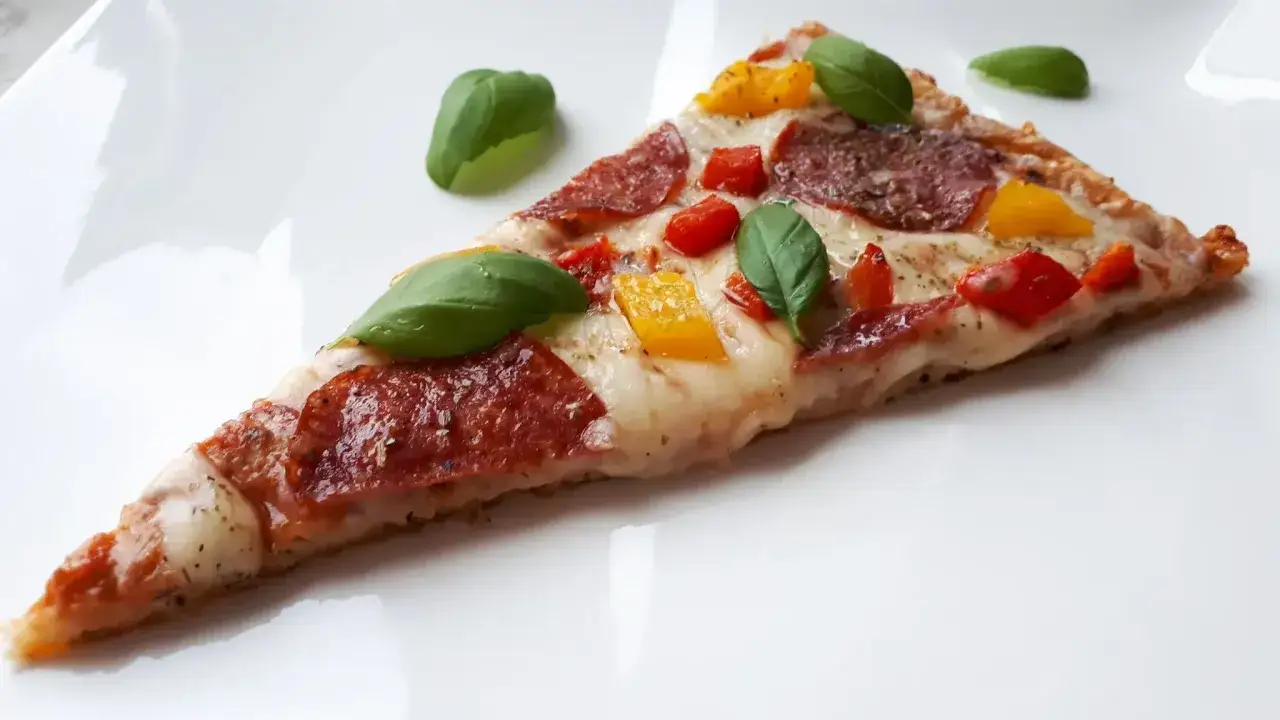 Kawałek pizzy z salami, papryką i bazylią na białym talerzu. Czy mozzarella jest keto? Tak, w umiarkowanych ilościach.
