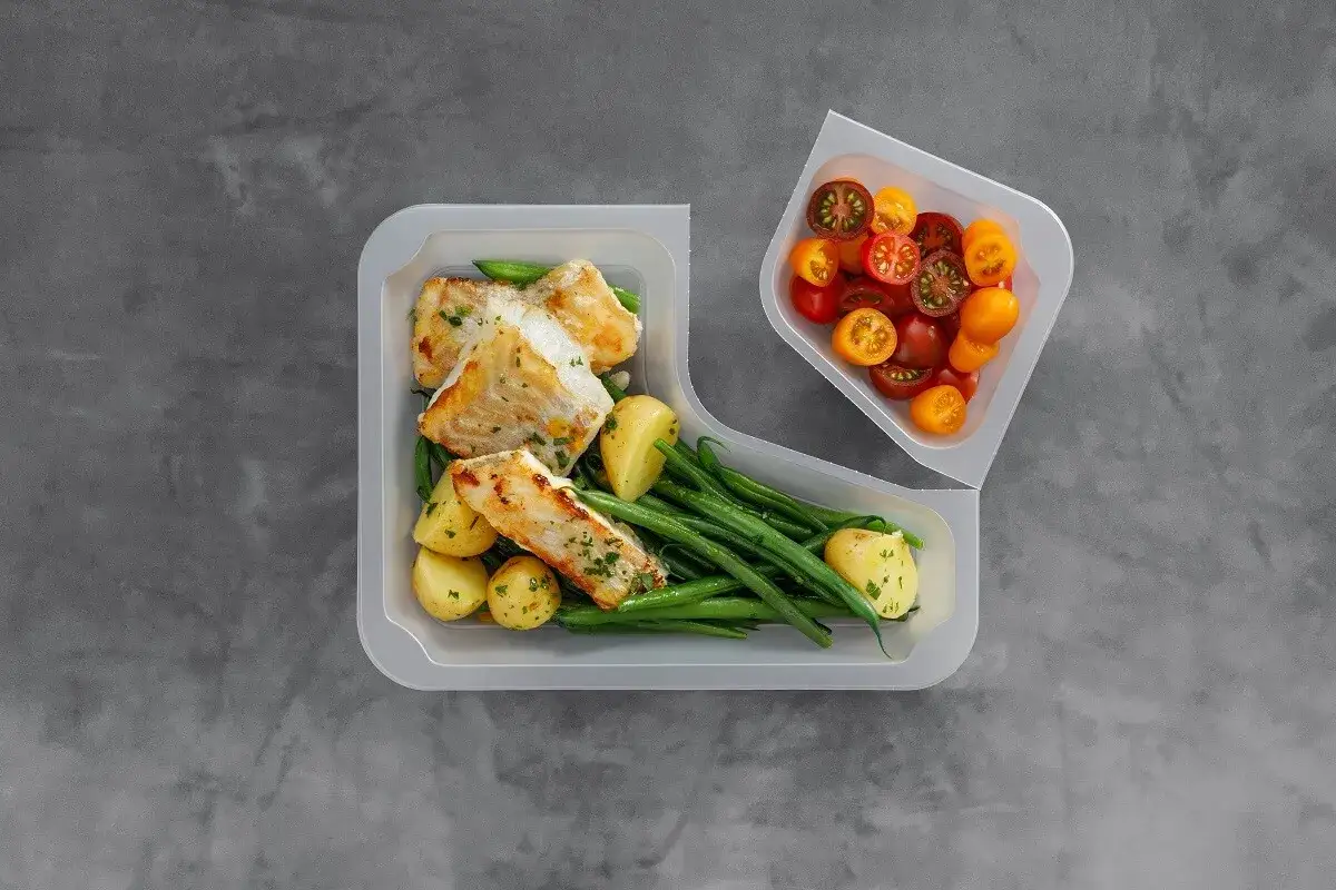 Jak podgrzewać jedzenie z cateringu? Danie z rybą, ziemniakami i fasolką szparagową w pojemniku, obok miseczka z pomidorkami.