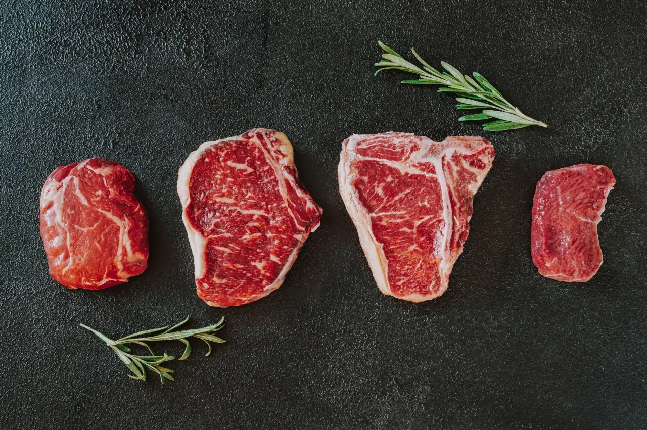 Różne kawałki wołowiny, w tym stek T-bone i polędwica, ułożone na czarnym tle z gałązkami rozmarynu. Idealne składniki, by dowiedzieć się, jak przyrządzać wołowinę.