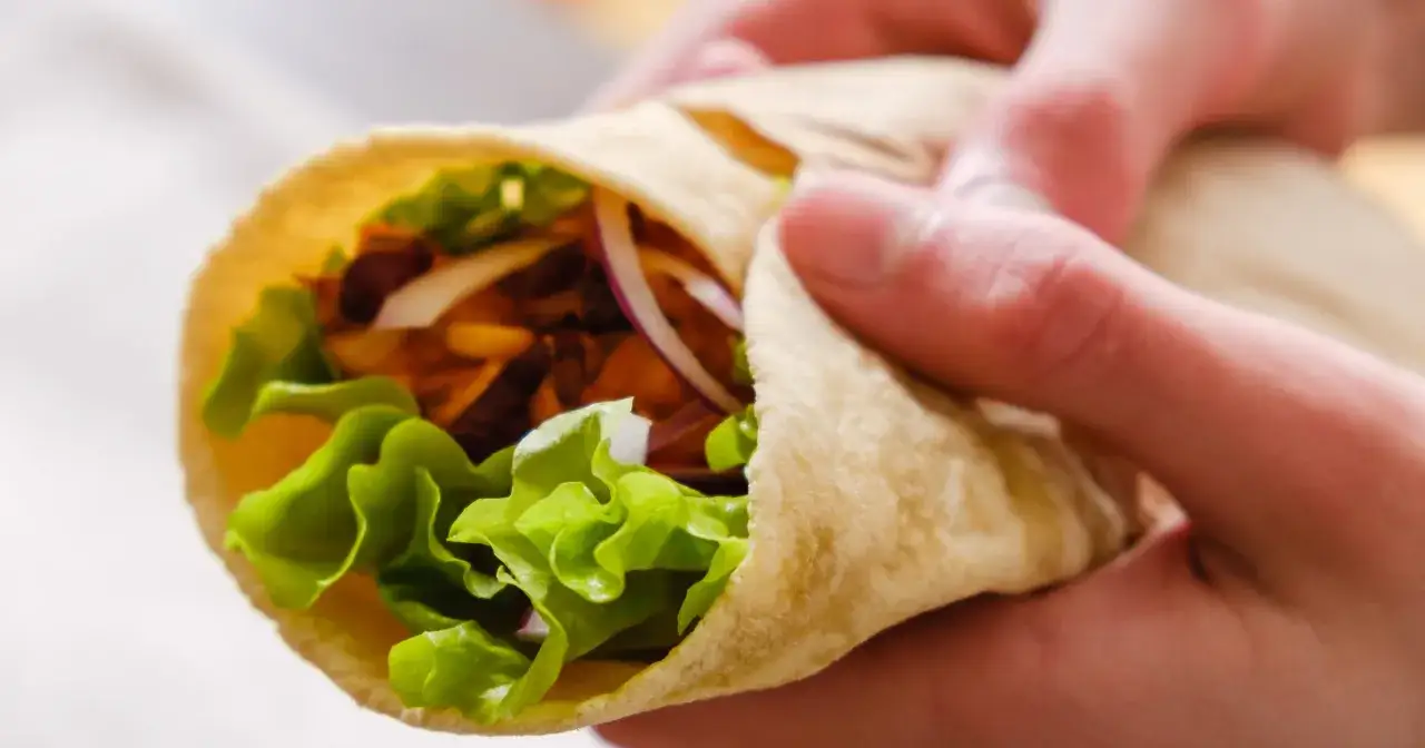 Ręce trzymają wrap z sałatą, cebulą i mięsem. Czy placki tortilli są zdrowe? Zależy od składników!