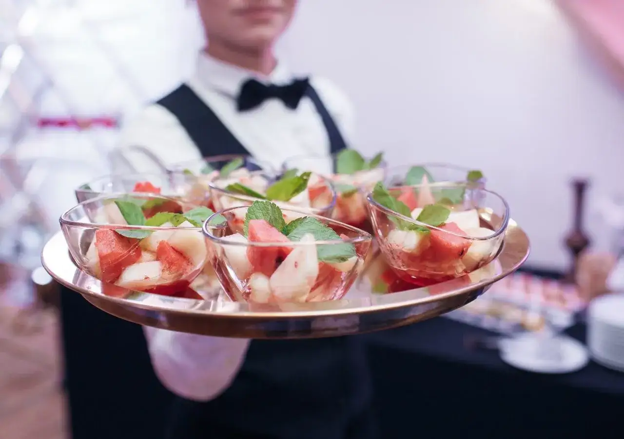 Kelner niesie tacę z owocowymi deserami w pucharkach. Zastanawiasz się, ile kosztuje catering firmowy?